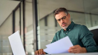 Busy middle aged executive, mature ceo, male hr account manager holding documents using laptop computer in office, checking financial bill, data in report, doing accounting paper file plan overview.