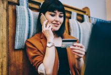 Positive woman looking at number on credit card and confirm purchase via telephone call to customer service, smiling hipster girl making payment via smartphone conversation while resting indoors