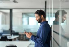 Busy Indian business man holding mobile phone device looking at cellphone standing at work. Professional businessman using finance banking apps on smartphone working on cell phone in office. Side view