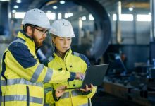 Male and Female Industrial Engineers in Hard Hats Discuss New Project while Using Laptop. They Make Showing Gestures.They Work in a Heavy Industry Manufacturing Factory.