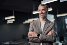 Happy confident mature 45 years old business man investor standing in office looking away. Middle aged rich business owner male ceo executive leader wearing suit at work thinking on future success.