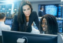 Female Team Leader Consults Young Computer Engineer. They Work in a Crowded Office on a Neural Network/ Artificial Intelligence Project.