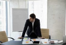 Frustrated concerned business man standing at table with sales reports, paper sticky notes, looking away, thinking over company future, negative statistics result, financial loss