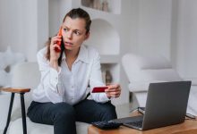 Worried businesswoman talks by phone with bank employee holds credit card sitting at desk with laptop tries to resolve problems, irritated by partners mistakes. Crisis, failure.