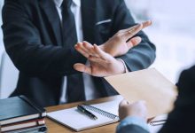 A businessman crosses his arms, forming an X, as another businessman tries to hand him an envelope.